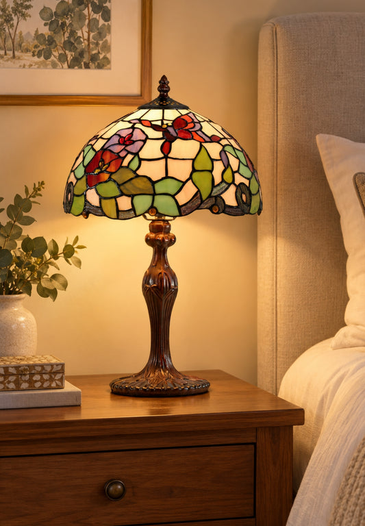A Tiffany-style stained glass table lamp with a floral pattern and amber-brown base, illuminated on a wooden bedside table next to a bed.