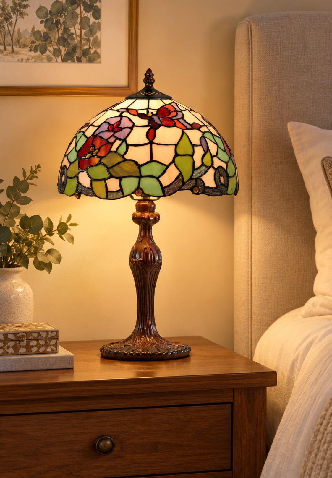 A Tiffany-style stained glass table lamp with a floral pattern and amber-brown base, illuminated on a wooden bedside table next to a bed.