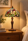 A Tiffany-style stained glass table lamp with a floral pattern and amber-brown base, illuminated on a wooden bedside table next to a bed.